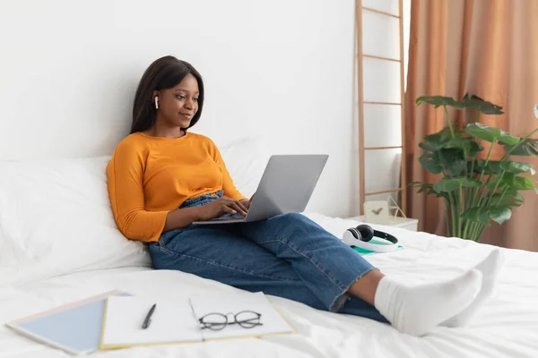 Mujer africana usando portátil usando auriculares trabajando en línea en el dormitorio — Foto de Stock