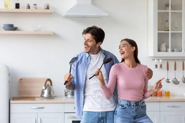Vidám fiatal kaukázusi pár énekelni képzeletbeli mikrofon, érezd jól magad, és táncolj a modern konyhában — Stock Fotó