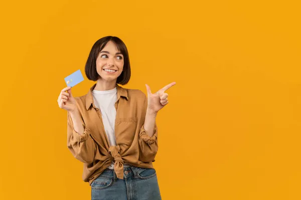 Nadšený kavkazský dáma s kreditní kartou ukazuje a dívá se stranou na prázdné místo na žlutém pozadí studia — Stock fotografie