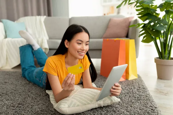 Ordinazione di merci online. Eccitato signora asiatica sdraiata su morbido tappeto con tablet pc e carta di credito, circondato da sacchetti regalo — Foto Stock