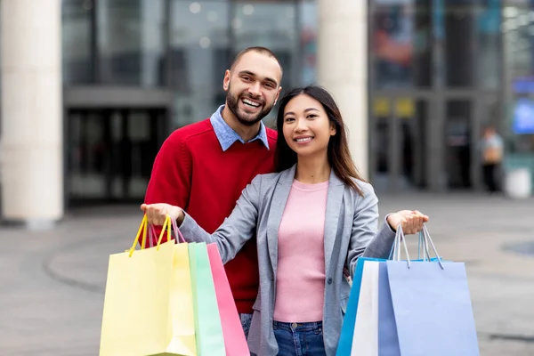 Atractiva pareja joven y diversa con coloridas bolsas de regalo mirando a la cámara y sonriendo cerca de un gran centro comercial —  Fotos de Stock