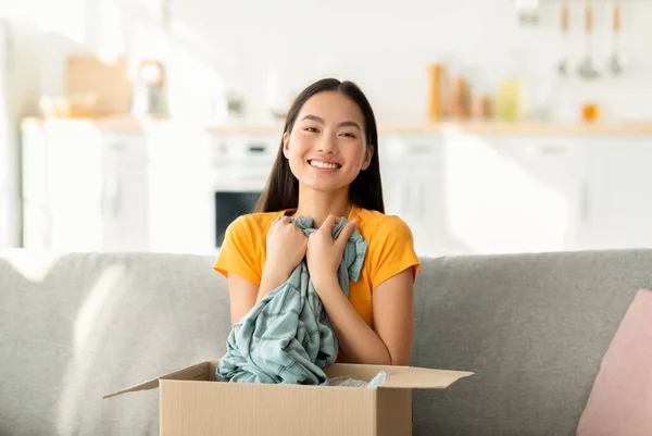 Zufriedenes Käufer-Konzept. Überglückliche asiatische Frau öffnet und entpackt Paket nach der Lieferung, umarmt Hemd — Stockfoto