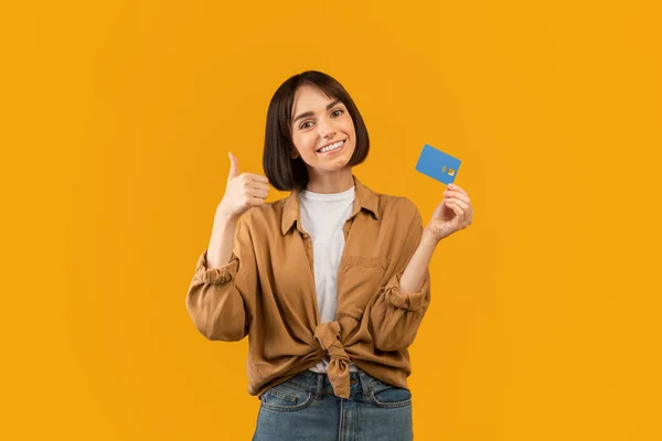 Весела молода леді показує кредитну картку і жестикулює великий палець, рекомендує банківське обслуговування, жовтий фон — стокове фото