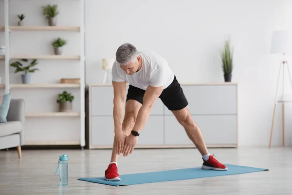 Hombre mayor deportivo estirando sus piernas, ejercitándose en casa, haciendo ejercicios de flexibilidad y llevando un estilo de vida saludable, de larga duración — Foto de Stock