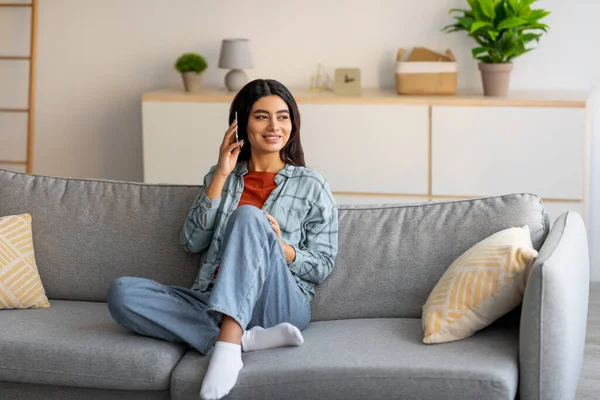 Jovem árabe feliz falando no smartphone, sentado no sofá em casa, espaço livre — Fotografia de Stock