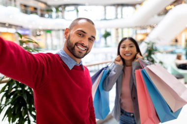 Mutlu uluslararası çift hediye çantalarıyla büyük alışveriş merkezlerinde selfie çekiyorlar, birlikte alışverişe gidiyorlar, fotokopi çekiyorlar.