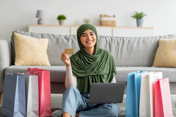 İnternet üzerinden alışveriş yapan, kredi kartı kullanan, kağıt torbalarla yerde oturan neşeli, tesettürlü genç Arap bayan. — Stok fotoğraf