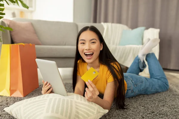 Ventas en línea y descuentos. Sorprendido asiático dama acostado en el suelo con tableta y tarjeta de crédito, emocionalmente reaccionando venta — Foto de Stock
