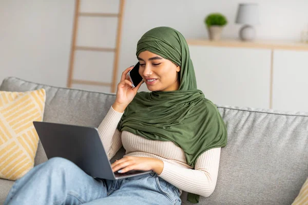 Retrato de una joven árabe usando una computadora portátil y hablando por teléfono celular mientras está sentada en el sofá en casa — Foto de Stock