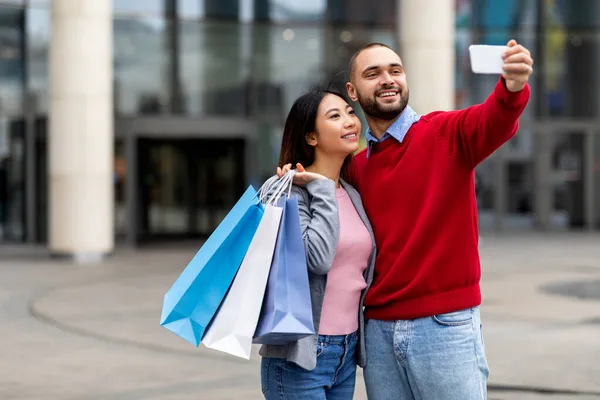 Portret młodej wielorasowej pary robiącej selfie na smartfonie, trzymającej jasne torby prezentów po zakupach w centrum handlowym — Zdjęcie stockowe