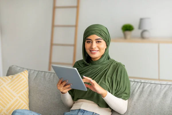 Başörtülü genç Arap kadın, evdeki rahat kanepede çalışmak, iletişim kurmak ve çalışmak için dijital tablet kullanıyor. — Stok fotoğraf