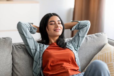 Rahat genç bir Arap kadın kanepeye yaslanıyor, elleri rahat koltuğun arkasında dinleniyor, gözlerini kapıyor.