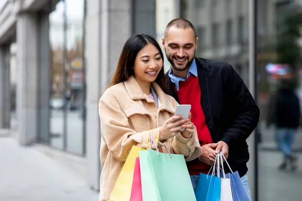 Positief jong divers koppel met heldere papieren tassen en mobiele telefoon om goederen online te bestellen in de buurt van winkelcentrum — Stockfoto