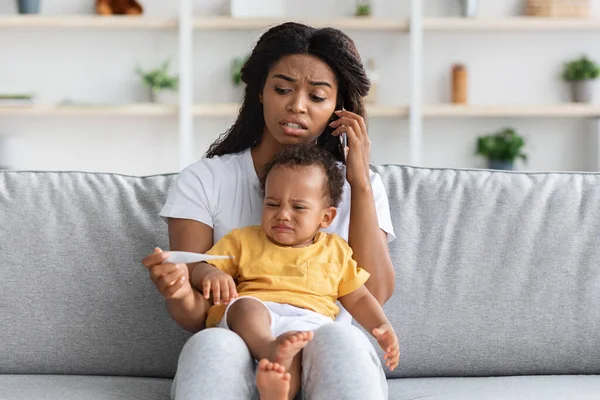 Bezorgde zwarte vrouw met kleine baby vasthouden thermometer en een beroep op dokter — Stockfoto