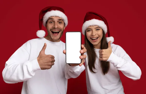 Mockup Image Of Happy Young Couple In Santa Hats Holding Blank Smartphone — Stock Photo, Image