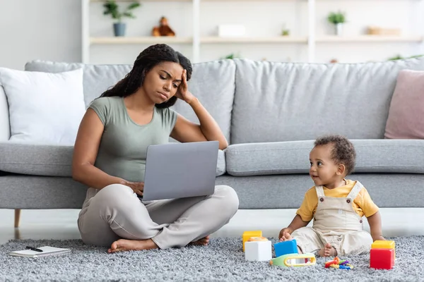 Виснажена чорна мати намагається працювати на ноутбуці з маленькою дитиною навколо — стокове фото