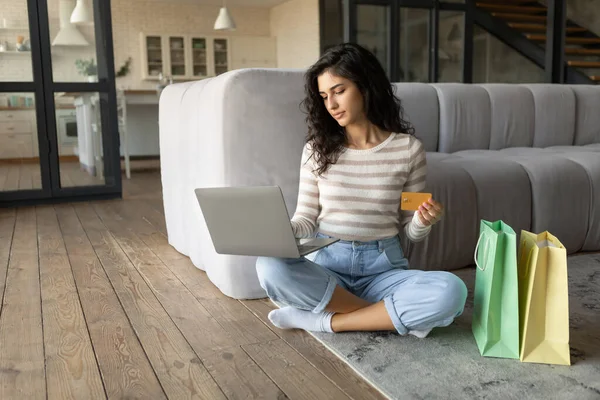 Feliz mulher milenar sentado no chão na sala de estar, compras on-line, segurando cartão de crédito e usando laptop em casa — Fotografia de Stock