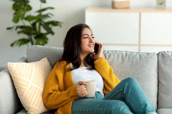 Ev içi rahatlık. Arap kadın cep telefonuyla konuşuyor ve evdeki kanepede dinlenirken kahve içiyor.