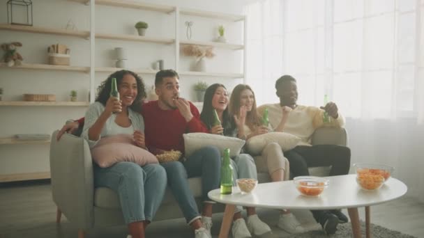 Группа молодых энтузиастов разнообразных друзей смотрят спортивный матч по телевизору, сидят на диване с пивом и закусками, увеличивают масштаб — стоковое видео