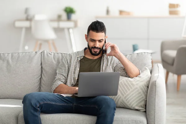 Schöner arabischer Millennial-Mann arbeitet am Laptop und telefoniert mit dem Smartphone, hat einen Job in der Ferne, drinnen — Stockfoto