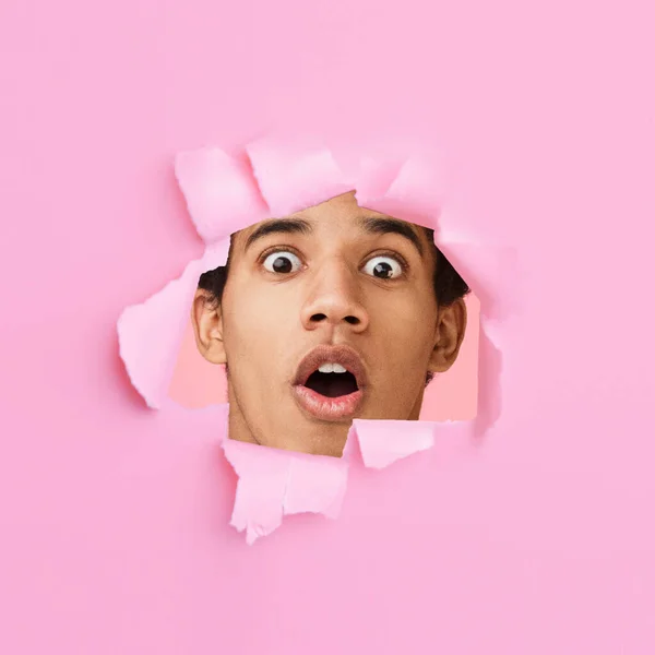 Scioccato stupito millenario ragazzo africano americano con bocca aperta guarda attraverso il buco in carta rosa — Foto Stock