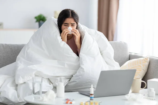 Frustration ung indian dam insvept i filt blåser näsa i servett på soffan med laptop och medicin — Stockfoto