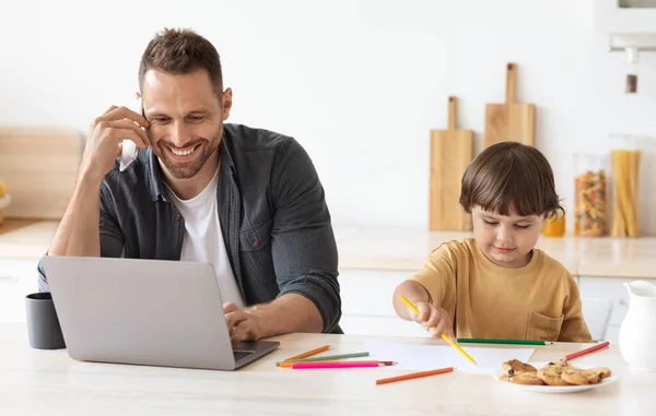 Cute little boy drawing pictures with colorful pencils, sitting at kitchen near working with laptop and cellphone father — Stock Photo, Image