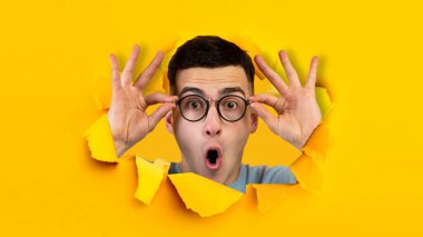 Ağzı açık, bardaklara dokunan, sarı kağıttaki delikten bakan pozitif Avrupalı bir genç.