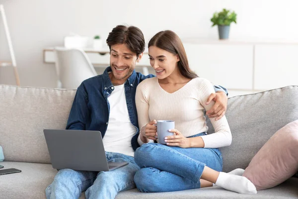 Chocante sorrindo milenar casal europeu sentado no sofá olhando para laptop, compras, chamando online — Fotografia de Stock