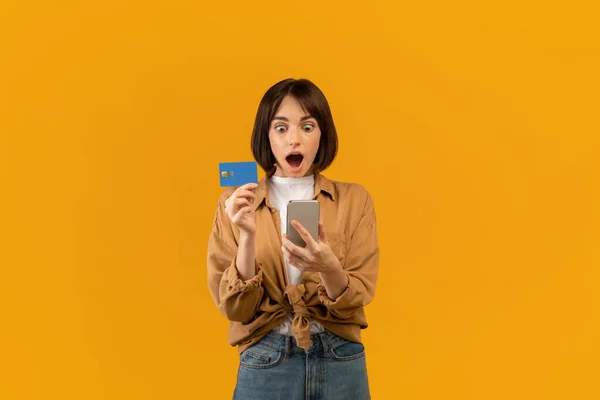 Концепція бонусу та кешбек. Здивована молода леді показує дебетову кредитну картку, використовуючи смартфон, роблячи онлайн-платежі — стокове фото