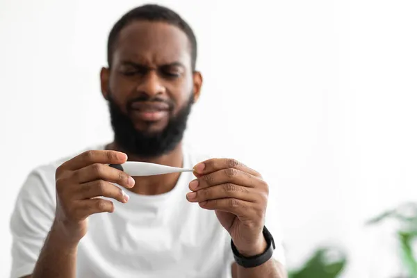 Triste joven afroamericano con barba mira el termómetro y se siente mal en el fondo blanco —  Fotos de Stock