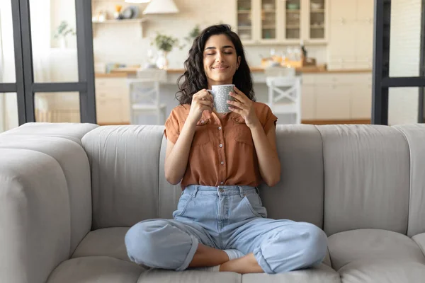 Bella giovane signora caucasica seduta a gambe incrociate sul divano, odore di caffè aromatico fresco con gli occhi chiusi a casa — Foto Stock