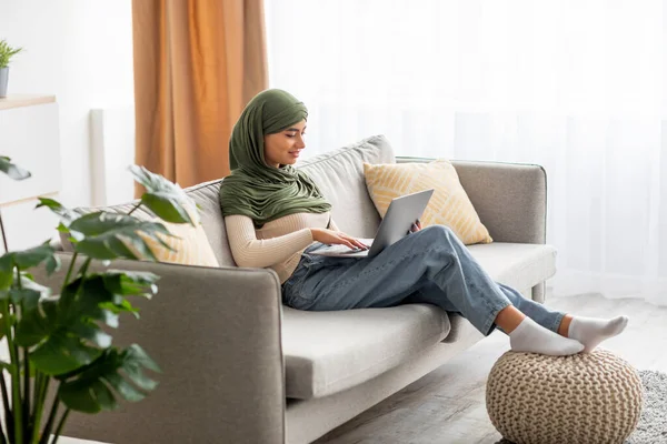 Mujer árabe joven en hijab sentado en el sofá, el uso de la computadora portátil, comprobar el correo electrónico, navegar por la web, trabajar o estudiar en línea en casa — Foto de Stock