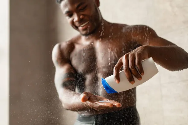 Yakışıklı siyah adam duş alıyor. Banyoda şampuan şişesi sıkıyor. — Stok fotoğraf