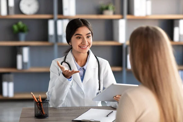 Sorridente giovane attraente indiano signora medico lavoratore in bianco cappotto gesti e consultazione paziente sul posto di lavoro — Foto Stock