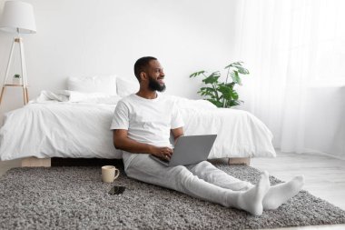 Mutlu genç siyah sakallı adam dizüstü bilgisayarda çalışıyor, yatak odasında iç tarafta oturuyor, pencereye bakıyor.