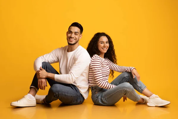 Joyeux jeune couple arabe assis dos à dos sur fond jaune studio — Photo