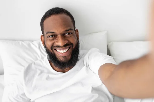 Zâmbind millennial african american barbă om se uită la webcam, se află pe pat alb în dormitor interior — Fotografie, imagine de stoc