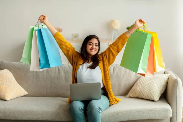 Alegre árabe señora de compras en línea, sentado en el sofá con el ordenador portátil, levantando bolsas de regalo por encima de su cabeza en la emoción — Foto de Stock