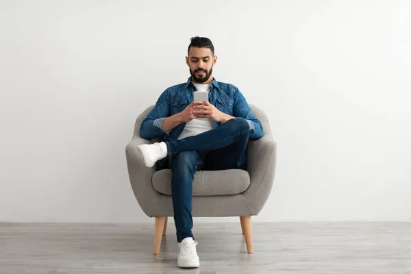Ung arabisk kille som använder mobiltelefon, chattar online, sitter i fåtölj, surfar sociala medier mot vit studiovägg — Stockfoto