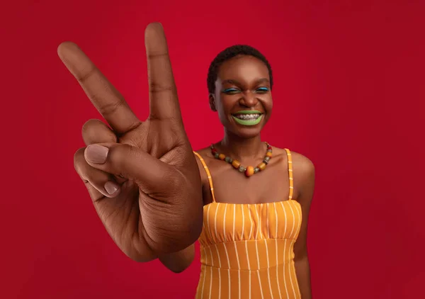 Heureuse femme noire avec un maquillage lumineux portant une tenue élégante, montrant une grande victoire ou un geste de paix sur fond rouge — Photo