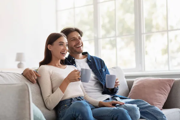 Sorrindo relaxado jovem europeu homem e mulher com xícaras de bebida quente desfrutar de tempo livre e assistir filme — Fotografia de Stock