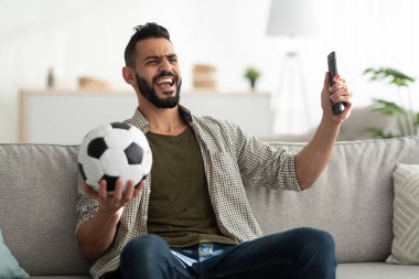 Televizyonda futbol maçı izleyen neşeli genç bir Arap adam elinde top ve uzaktan kumandayla evdeki kanepede oturuyor.