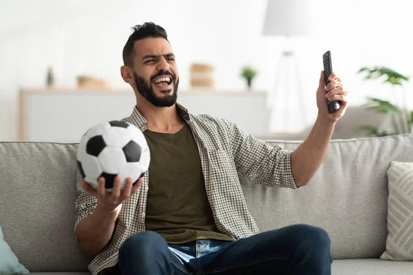 Fröhlicher junger Araber schaut Fußballspiel im Fernsehen, hält Ball und Fernbedienung in der Hand, sitzt zu Hause auf der Couch — Stockfoto