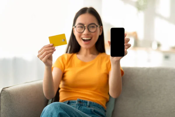 Banking, purchasing concept. Excited asian woman showing blank smartphone screen and credit card, sitting on sofa — Stock Photo, Image