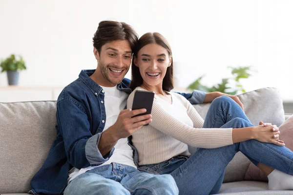 Sonriendo sorprendido joven caucásico marido y mujer mira en el teléfono inteligente, tener reunión en la sala de estar —  Fotos de Stock