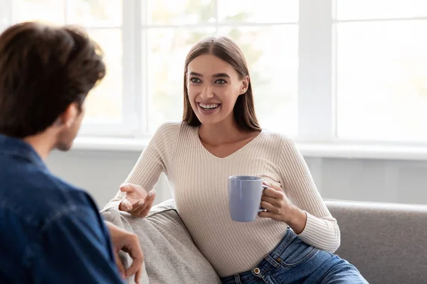 Vidám, fiatal európai férj és nő egy csésze forró itallal beszélgetni és együtt tölteni a hétvégét — Stock Fotó