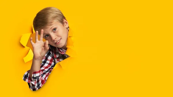 Vidám európai tinédzser szőke fiú mutatja rendben jel, néz át lyukon sárga papír, panoráma — Stock Fotó