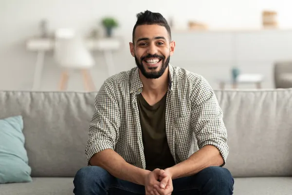 一个英俊的阿拉伯年轻人面带微笑地看着相机，坐在家里沙发上的画像 — 图库照片