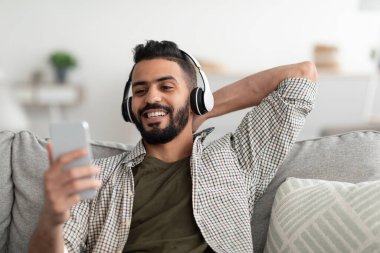 Rahatlama zamanı. Kablosuz kulaklıklı yakışıklı genç Arap evde akıllı telefondan müzik dinliyor.